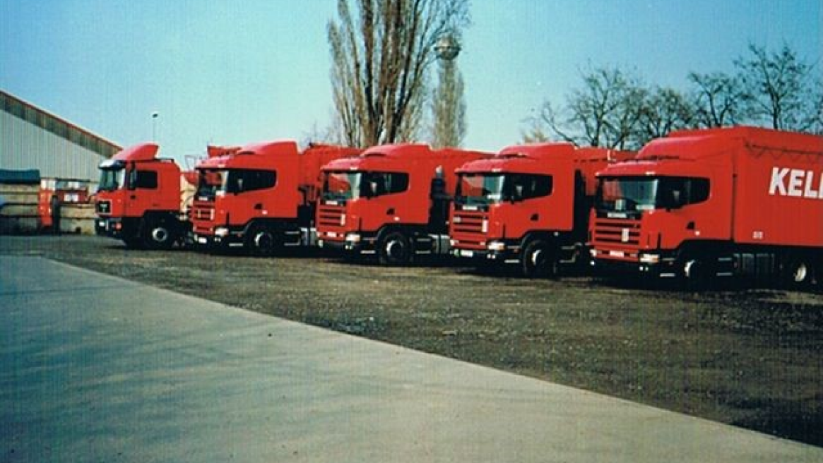Eine Reihe von roten LKWs mit dem Aufdruck ‘KELLER’ geparkt auf einem offenen Gelände mit Bäumen und einem Gebäude im Hintergrund.