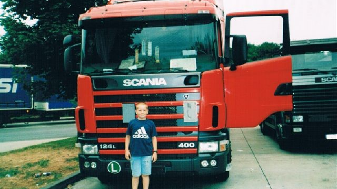 Ein Bild eines roten SCANIA-LKWs mit einem Kind davor, und einem weiteren LKW im Hintergrund.