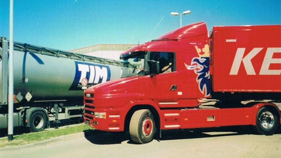Ein roter LKW mit dem Scania Logo, der neben einem Tankwagen mit dem Logo ‘TIM’ parkt, unter einem klaren blauen Himmel.