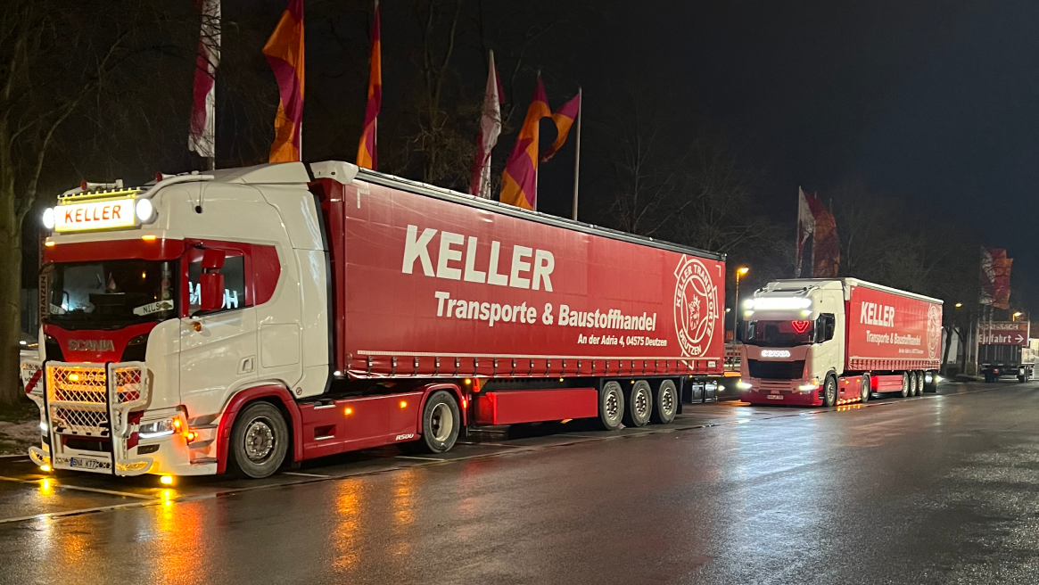 Zwei beleuchtete Keller Transporte & Baustoffhandel LKWs in der Nacht, geparkt auf einer nassen Straße mit Flaggen im Hintergrund.